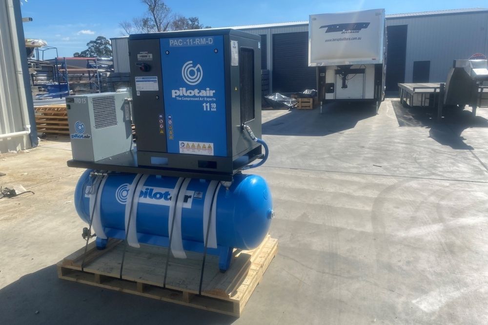 A large blue Pilot Air industrial air compressor unit sits secured on a wooden pallet in an outdoor works